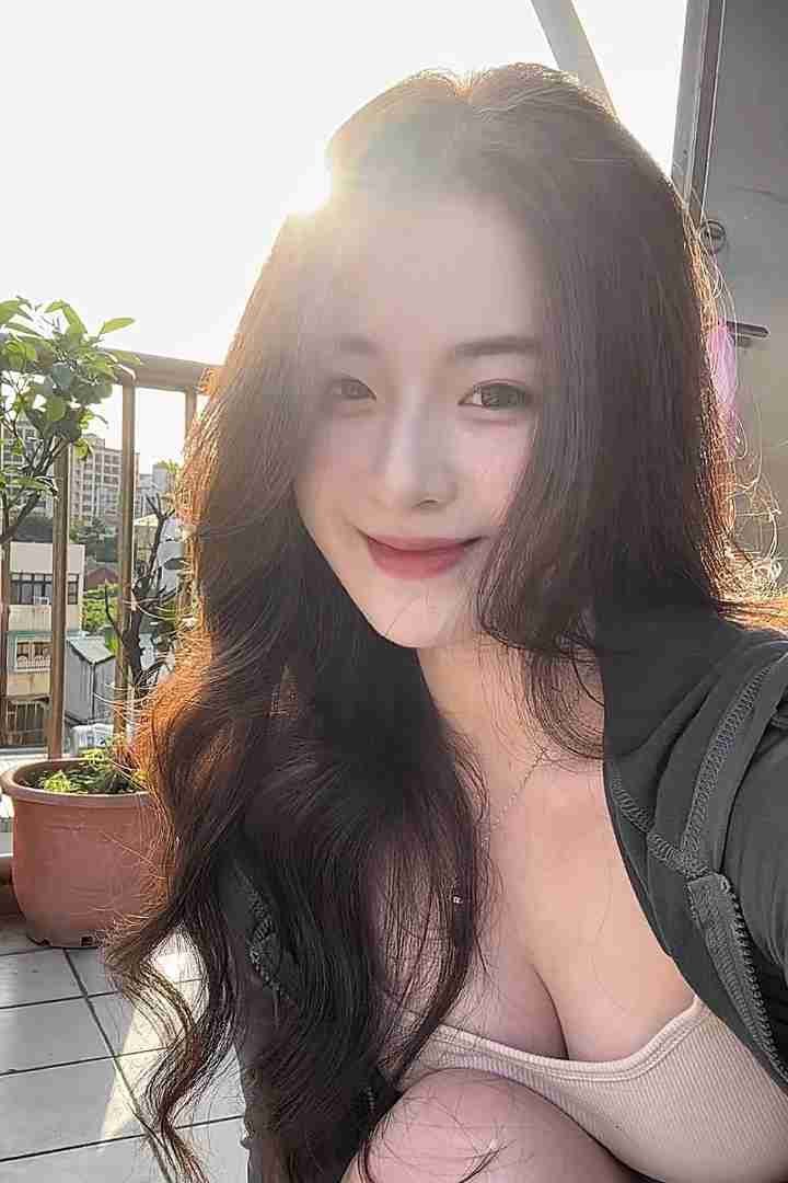 KL Escort Girl Smiling woman with long, dark wavy hair posing on a balcony with city buildings in the background. Sex Call Girl