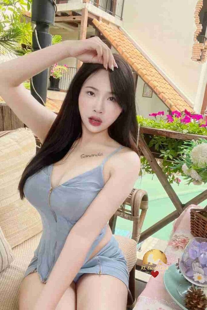 Asian woman in blue top posing outdoors with flowers. Tattoo on chest says "Sananda".