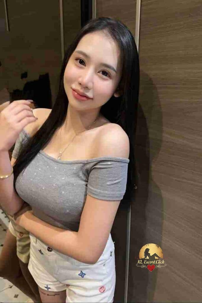 Smiling Asian woman in gray top and white shorts. Long black hair, gold necklace, and bracelet.