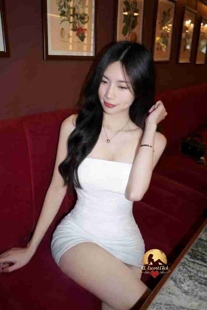 Elegant Asian woman in white dress sitting on red couch. Black hair, necklace, stylish pose. Interior setting.