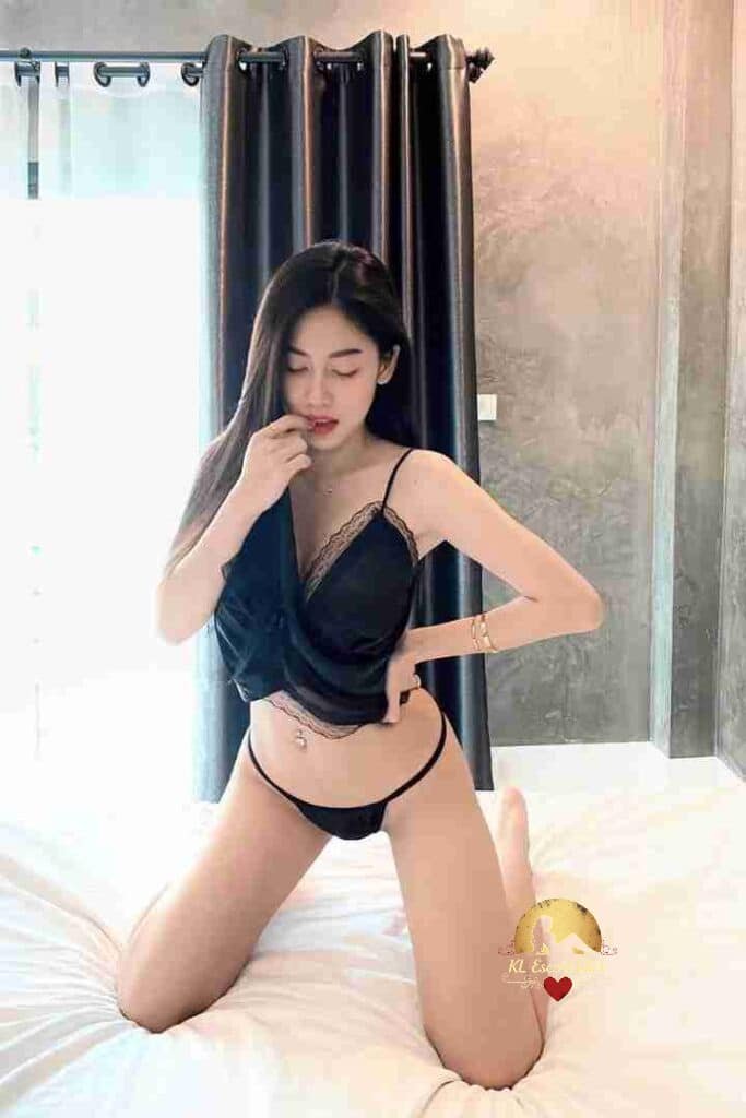 Woman in lingerie kneeling on bed, hand to mouth. Black top and bottom set, bedroom setting.