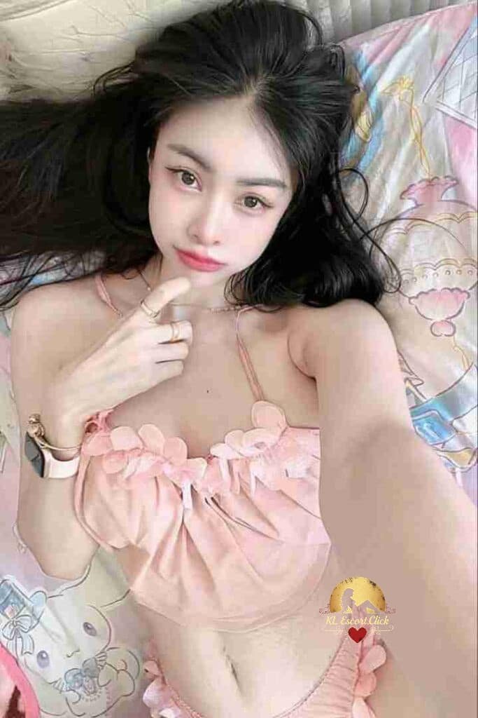 Woman in pink lingerie lying on patterned bed. Black hair, pink lips, delicate jewelry, and a watch are visible.