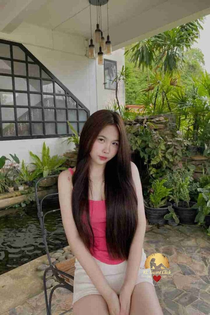 Young woman with long brown hair in pink top and white shorts sitting outside near plants and a pond.